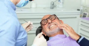 Older male patient in a dental chair wincing and holding his jaw during an exam, highlighting dental discomfort or tmj issues often seen in senior oral health care.