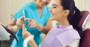 Smiling female patient looking at her teeth in a handheld mirror while a dental hygienist in scrubs provides guidance, illustrating a positive preventive care visit.