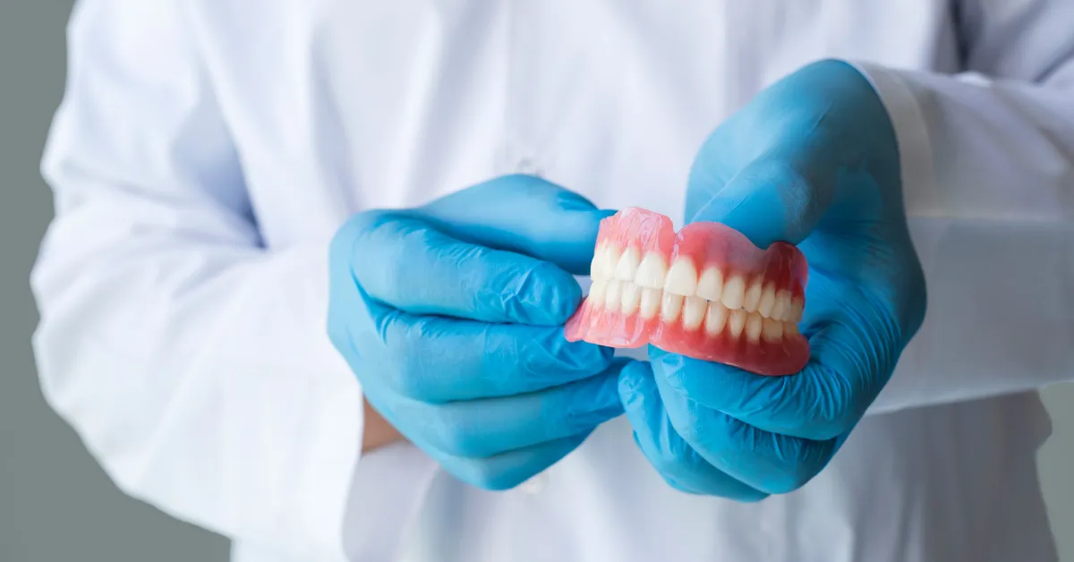 Dentist wearing blue gloves holding a full set of upper and lower dentures, representing professional dental prosthetics and restorative oral care.